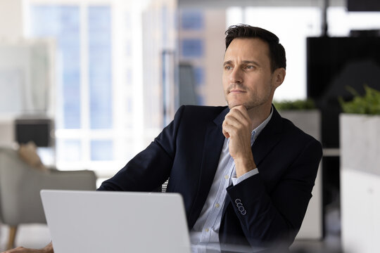 Serious Hispanic middle-aged man dressed in suit sit at desk with open laptop looks away with pensive, thoughtful expression, ponder on issue solution or new idea, strategizing in contemporary office - Powered by Adobe