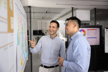 Two men stand in front of large whiteboard covered with sticky notes and detailed workflow plan, smiling, pointing at board engaged in discussion, productive, positive collaboration in modern office