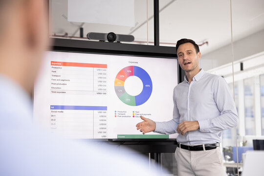 Businessman, financial analyst makes presentations, standing next to large screen displaying colorful pie charts representing various expenses categories, provide explanations share strategic decision