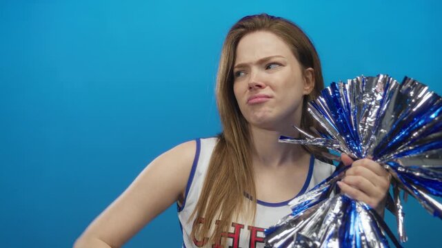 Woman in white cheer uniform holds metallic blue pompom and touches chin in a blue studio; doubt thought pondering.