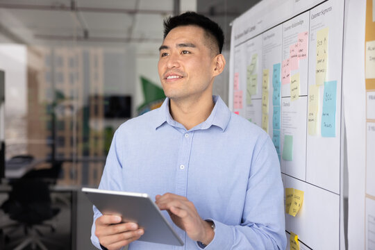 Project development, innovation, planning and strategic thinking. Young 30s man holding tablet and looking thoughtfully away with pensive expression stands next to whiteboard covered with sticky notes