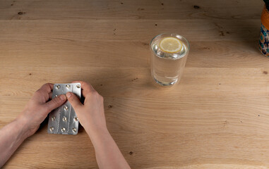 A pair of hands holding a silver blister pack filled with pills, poised above a light wooden table, with a glass of water containing a lemon slice in the background, ready for medication
