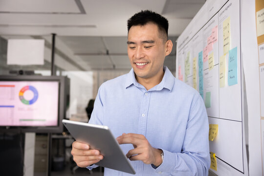 Chinese businessman taking digital notes or making edits to project plan using modern tablet device, reviewing new software, learns lists tasks to-do through corporate application working in office