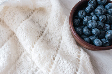 Fresh blueberries in clay bowl and white knitting. Knitted pattern and cotton knitted cardigan. Summer berries and cotton knitted sweater. Summer still life. Organic food and knitting hobby. 