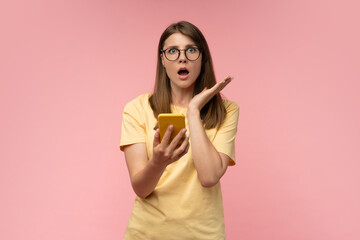 Shocked young woman in trendy glasses looking at camera with surprised face, holding smartphone in...