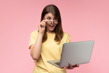 Studio shot of a happy young casually dressed woman holding using laptop computer and looking at its screen with excited face expression, isolated over pastel pink background