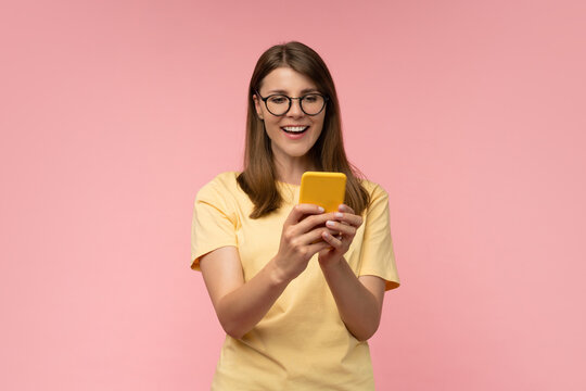 Smiling young woman in trendy glasses scrolling smartphone on pastel pink background