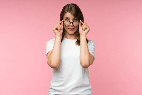 Portrait of attractive young woman posing touching specs with surprised face expression, isolated over pastel pink color background