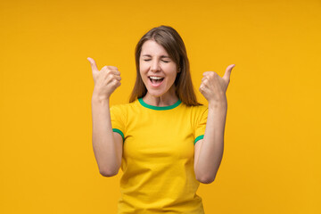 Excited young woman in yellow shirt celebrating success with double thumbs up on yellow studio...