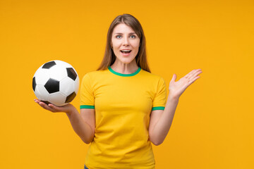 Young woman on yellow studio background holding a soccer ball with surprised and excited wow open mouth face expression, showing excitement and passion as an athlete or fan