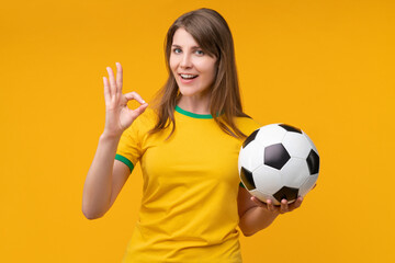 Young woman on yellow studio background holding a soccer ball and showing an OK hand gesture, expressing confidence, positivity, and support as a cheerful fan or athlete