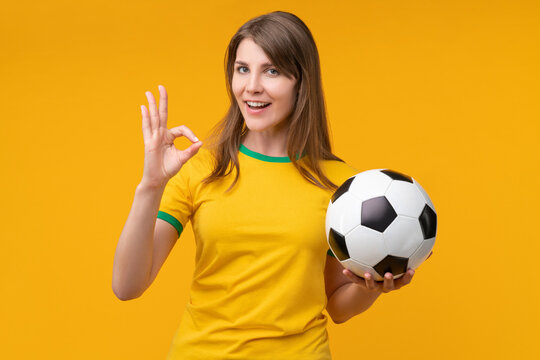 Young woman on yellow studio background holding a soccer ball and showing an OK hand gesture, expressing confidence, positivity, and support as a cheerful fan or athlete