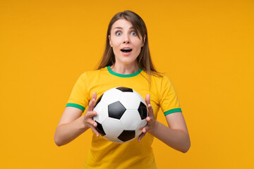 Young woman supporter in yellow t-shirt holding soccer ball with amazed face expression after catching it