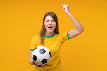 Confident young woman holding a soccer ball and raising fist in air in winners gesture while...