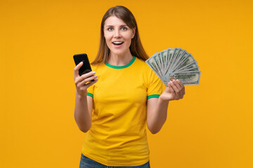 Happy young woman holding smartphone and fan of dollar bills, looking amazed