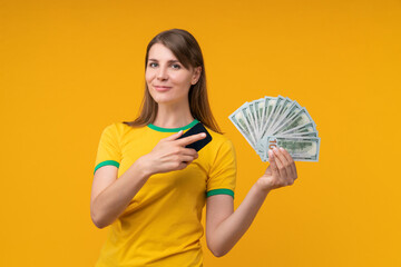 Smiling young woman in yellow shirt posing on yellow background, holding a smartphone and a fan of dollar bills, celebrating a money win from online betting or gamblin