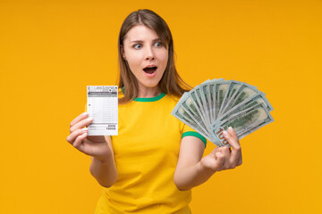 Shocked young woman posing on yellow background, holding a betting slip in one hand and staring at...