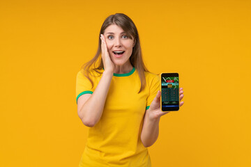 Excited young woman in yellow t-shirt holding smartphone with live betting screen, expressing...