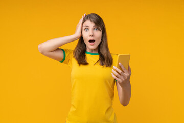 Young woman on yellow studio background holding a smartphone in one hand and clutching her head...