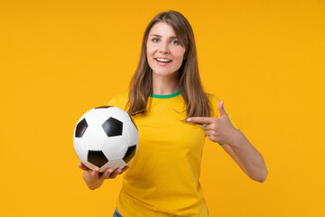 Young woman on yellow studio background holding soccer ball in one hand and pointing at it with the other, symbolizing passion for the game, sports betting, and the thrill of victory
