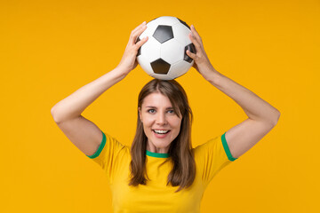 Young woman on yellow studio background holding soccer ball above her head, celebrating the spirit...