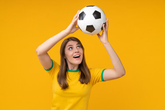 Joyful young woman in yellow t-shirt holding soccer ball above her head, looking at it with excitement - Powered by Adobe