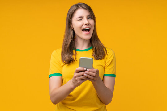 Young woman on yellow studio background winking while holding a smartphone, expressing confidence and playful excitement, suggesting success in betting, gambling, or winning money online - Powered by Adobe