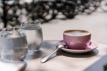 Minimal luxury aesthetic concept. Bohemian crystal glasses and pink cup of coffee with sparkling sunlight shadow on the table in a street cafe