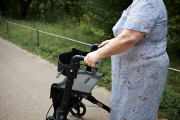 Obraz premium Elderly lady with walker enjoying a stroll in the park. Older lady with mobility aid walking along the street. Senior woman outdoors using a wheeled walker. 