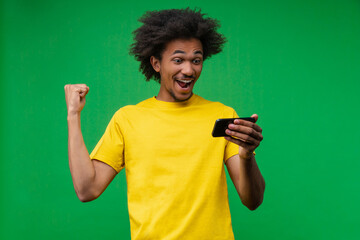Excited young African-American man on green background looking at smartphone screen and making a...