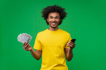 Smiling young African-American man looking directly at camera while posing with a smartphone in one hand and a fan of dollar bills in the other. Studio shot. Concept of betting and money win