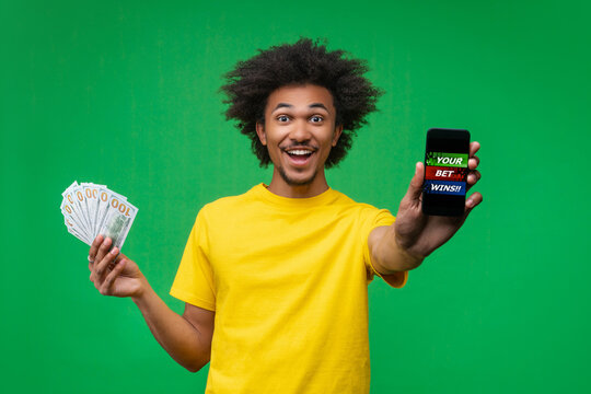 Confident young African-American man posing on green studio background, holding a fan of dollar bills and showing smartphone screen with a sample betting app design. Concept of gambling and money win
