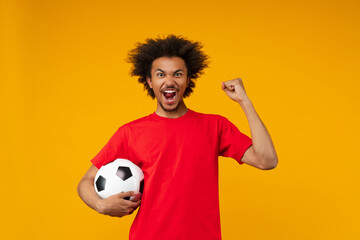 Young excited African American man poses on yellow studio background, holding a soccer ball in one...