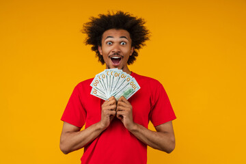 Young African American man poses on yellow studio background, holding a fan of dollar bills with a...