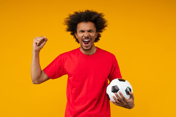 Excited African man on yellow background screaming with rage and passion while holding soccer ball...