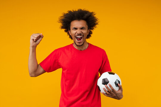 Excited African man on yellow background screaming with rage and passion while holding soccer ball and making a yes gesture