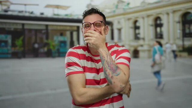 Young man smiling and gesturing excitedly in a vibrant urban street setting, showcasing colorful tattoos and wearing glasses and a striped shirt.