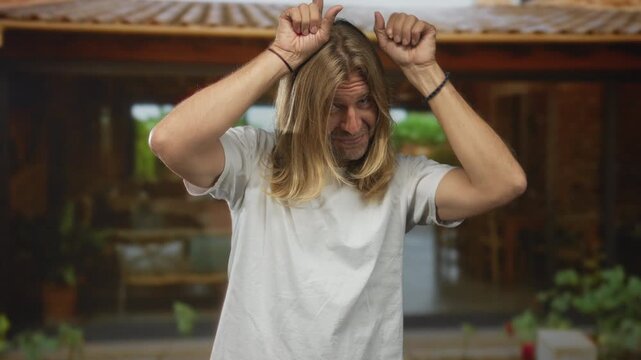 Blond man in white tshirt with long hair makes rock horns gesture at building entrance; rock metal fun.