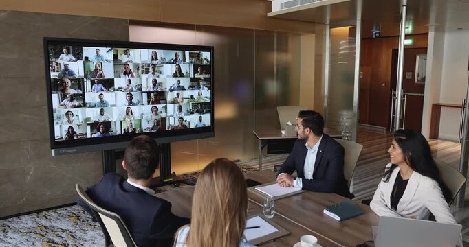 Multiethnic team of young businesspeople holding video conference meeting with many coworkers, partners, clients, sitting at large office table, looking at wall monitor with split head shots
