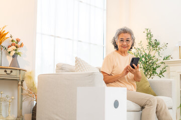 Happy Asian senior woman enjoy smart home system using smartphone app controlling electric appliance gadget device. Elderly woman relaxing on sofa using mobile phone turn on lighting and Air purifier.
