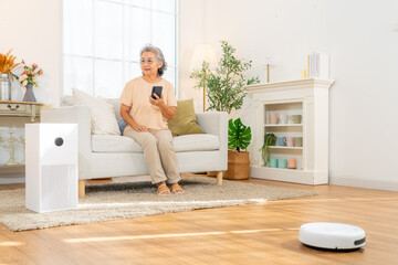 Happy Asian senior woman enjoy smart home system using smartphone app controlling electric appliance gadget device. Elderly woman relaxing on sofa and cleaning in living room with robotic vacuum.