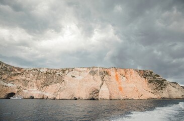 A tour boat approaches a rocky coastline in Zakynthos, Greece, on an overcast day. The cliffs rise...