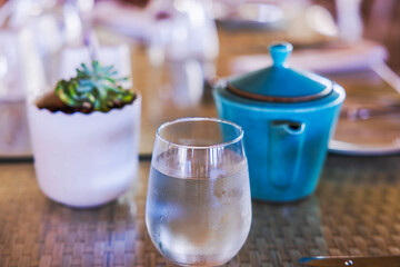 Close up view of glass of cold water with condensation near blue ceramic teapot and small plant on glass table. Dominican Republic. Punta Cana.