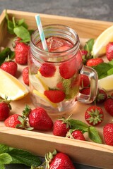 Refreshing drink with strawberries, lemon and mint in mason jar surrounded by fruits in wooden crate on grey table, closeup