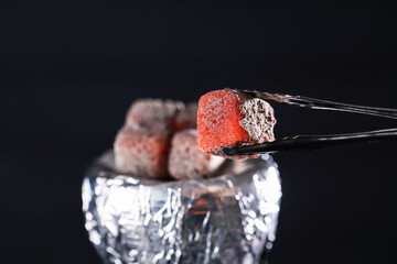 Holding smoldering charcoal cube with tongs on dark background, closeup