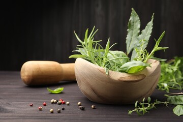 Mortar with different fresh herbs, peppercorns and pestle on wooden table against dark background,...