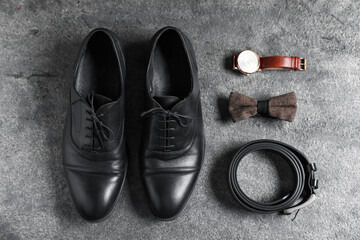 Pair of black leather shoes, wrist watch, belt and bow tie on grey surface, flat lay