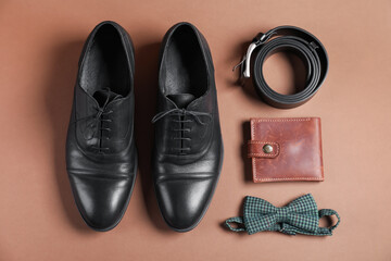 Pair of black leather shoes, belt, bow tie and purse on dark beige background, flat lay