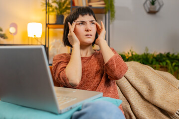 Tired young woman freelancer sitting on sofa using laptop, suffering from headache problem tension and migraine, stress. Caucasian adult girl feeling bored stretching arms sitting on couch at home.