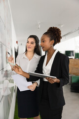 Two multiethnic women colleagues stands next to large white board holding tablet and papers, check...